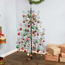 A metal Christmas ornament tree is decorated with red, green, and gold ornaments and displayed in a living room.
